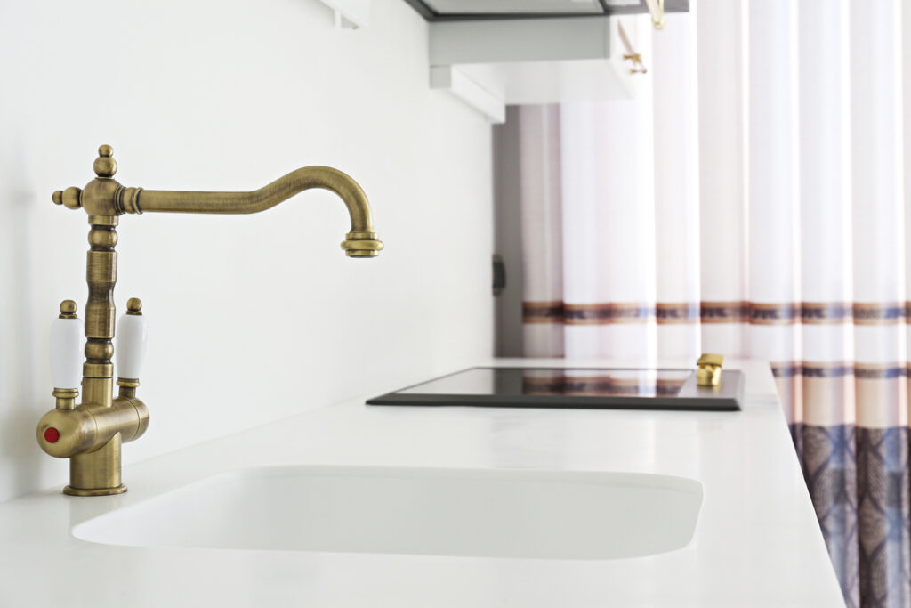 Faucet as the focus point. Image: brass-coloured vintage two-handle faucet on a white marble kitchen sink and stove range on the back