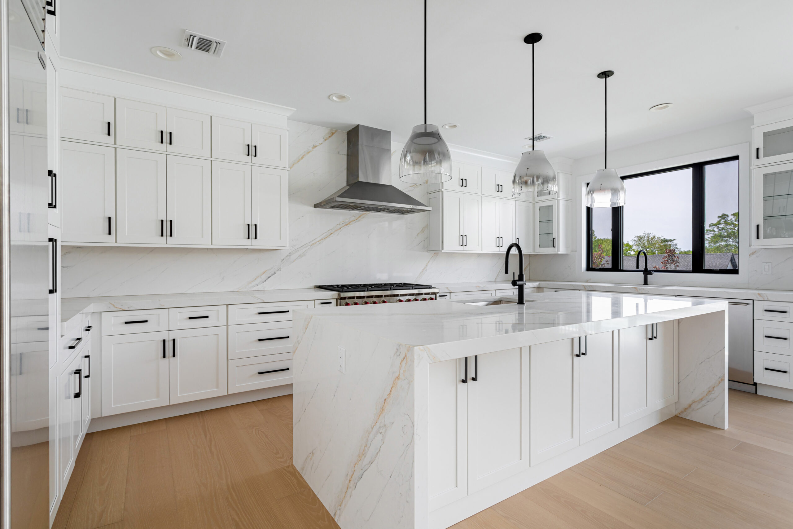 White modern kitchen with a large marble island, pendant lights, and a stainless steel range hood