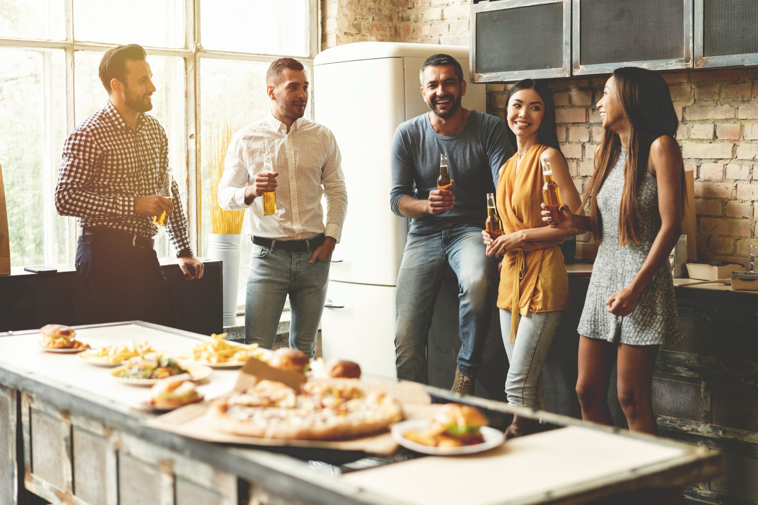 Friends chatting with drinks around a kitchen island set with pizza and snacks during a home gathering